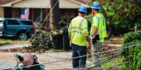 Two workers wearing hardhats survey damage on a street near a fallen electrical pole