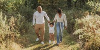 A dad, son, and mother hold hands and walk down a grassy path lined with bushes