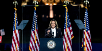 Kamala Harris speaks behind podium on stage behind a panel of bullet proof glass