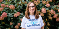 Sarah McBride stands in front of a bush with pink flowers and smiles