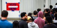 People line up to vote, a "Vote Here Sign" is taped to a wall nearby