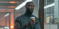 Aldis Hodge leans on a door frame in an office and holds a coffee