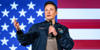 Elon Musk speaks on stage, with an American flag backdrop behind him