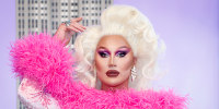 The Vivienne poses for a photo wearing a pink velvet dress and blonde curly hair