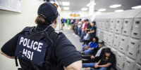 An officer stands in front of people seated and standing against lockers in a hallway