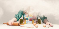 A woman in an apron, face obscured by a cloud of flour, sits at a table with a rolling pin and eggs