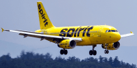 A yellow Spirit Airlines plane flies in the sky above trees