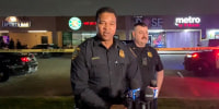 J. Skelton speaks into microphones in a parking lot with police cars and police tape, in front of another officer