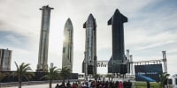 A crowd stands on a lawn in front of a stage, four large spaceship prototype sculptures in the background