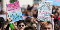 Two people hold signs outside in a crowd, sign on the left says "Trans women are real women", sign on the right says "Science says trans people are real"