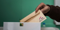 A hand places a beige ballot envelope into a white container