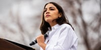 Alexandra Ocasio-Cortez holds a microphone to her chest while standing at a podium outside