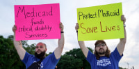Two people hold signs outside, one the left it says "Medicaid funds critical disability services", and on the right it says "Protect Medicaid, Save lives"