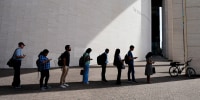 Eight people stand in a shadow to protect themselves from the sun.
