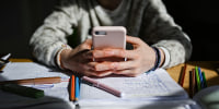 A child's hands hold a cellphone on top of a desk with homework and pens