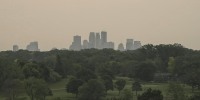 A hazy light orange sky seen above a skyline of buildings, trees are seen in the foreground