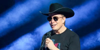 Elon Musk speaks onstage wearing a black cowboy hat, in front of two Tesla vehicles