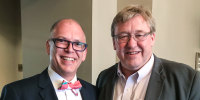 Jim Obergefell, left, and Rick Hodges stand together for a photo in a lobby, Jim holds a wooden letter V between them