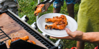 Outside by a lake, a man takes chicken drumsticks off of a grill onto a plate, kids surround him