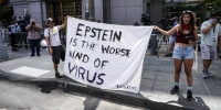 Two people hold a white blanket sign with black text that reads "Epstein is the worst kind of virus"