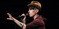 Andrea Gibson speaks into a microphone while standing on stage, one arm outstretched