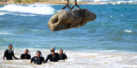 Divers watch as a crane pulls a piece of stone from the sea, with a cityscape in the background