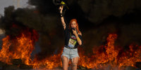 An activist blocks a highway during a protest, standing in front of a fire.
