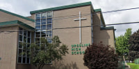 The exterior of Ursuline High School, a large white cross hangs on the beige brick side of the building on top of green text that reads "Ursuline High School"