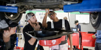 Three people look underneath a car that is lifted up inside of a classroom