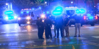 Police stand outside on the street in front of police cars with lights on, police tape is in front of them
