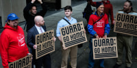 People stand on the street outside holding brown cardboard signs that say "Guard: Go Home!" in black text