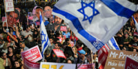 A crowd of protestors, some people wave large Israeli flags and posters