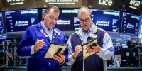 Two people look at tablets on the floor of the New York Stock Exchange building, tv screens behind them