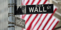A street sign that shows the intersection of Wall Street and Broad Street, American flags are seen blurred in the background
