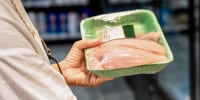 A woman in a white shirt shops in a supermarket, looking at products in a refrigerated section.
