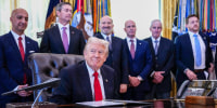 President Trump sits at his desk at the White House surrounded by others.