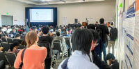 People stand and sit inside of a conference room, with a large projector screen showing a slideshow