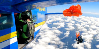 As seen from the side of a plae in the air, someone sits by the open door as another parachutist is in the air with their parachute stuck to a wing