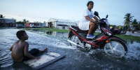 A woman rides her scooter through floodwaters next to a boy sitting near a plank on the flooded street