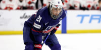 Hilary Knight stoops while holding a hockey stick on the ice. She is wearing a blue, red and white uniform and a white helmet.