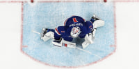 An overhead view of a goalie on the ice, in front of a puck that has drifted past the goal line and into the net.