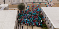 A dense crowd of hundreds of people wearing raincoats and hoods is seen from an aerial perspective. Many of them are holding signs.