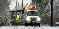 Two workers in blaze yellow suits work to clear fallen trees from a snow-covered roadway. 