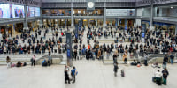 A crowd of people fills a large train hall with a glass roof, seen from an elevated perspective.