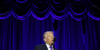 President Trump speaking into a microphone in front of a blue velvet stage backdrop.
