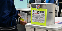 A man holding a voting ballot passes in front of a metal lockbox with a label reading DROP BOX FOR VOTED MAIL BALLOTS inside a polling station.