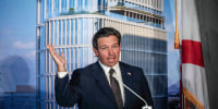 Ron DeSantis gestures with an open hand in front of a backdrop depicting a high rise office building.