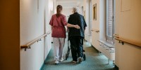 Rear view of female caregiver assisting senior woman walking with mobility walker in corridor