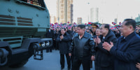 North Korean leader Kim Jong Un amongst a group of men while looking at appears to be a military truck. People stands behind holding North Korea's flag.