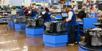 Cashiers and customers are lined up at a series of blue checkout aisles inside a Walmart. 